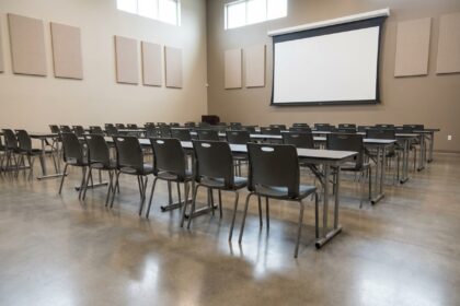 Salle polyvalente d'établissement configurée en disposition conférence avec rangées de chaises empilables et tables pliantes sur sol béton poli